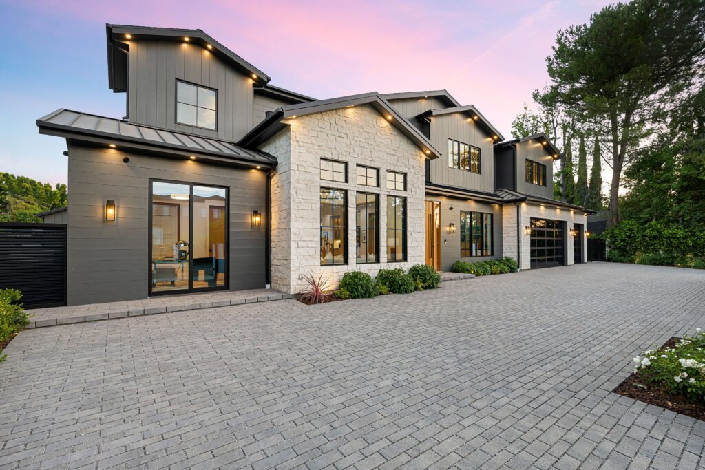 Front view of a modern two-story house with gray siding, stone accents, large windows, and a paved driveway at dusk.