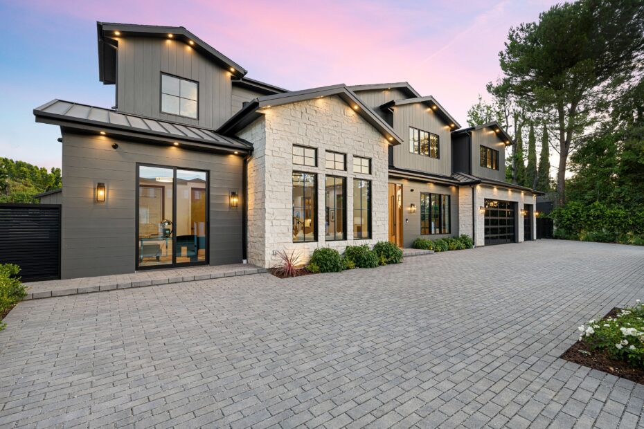 Front view of a modern two-story house with gray siding, stone accents, large windows, and a paved driveway at dusk.